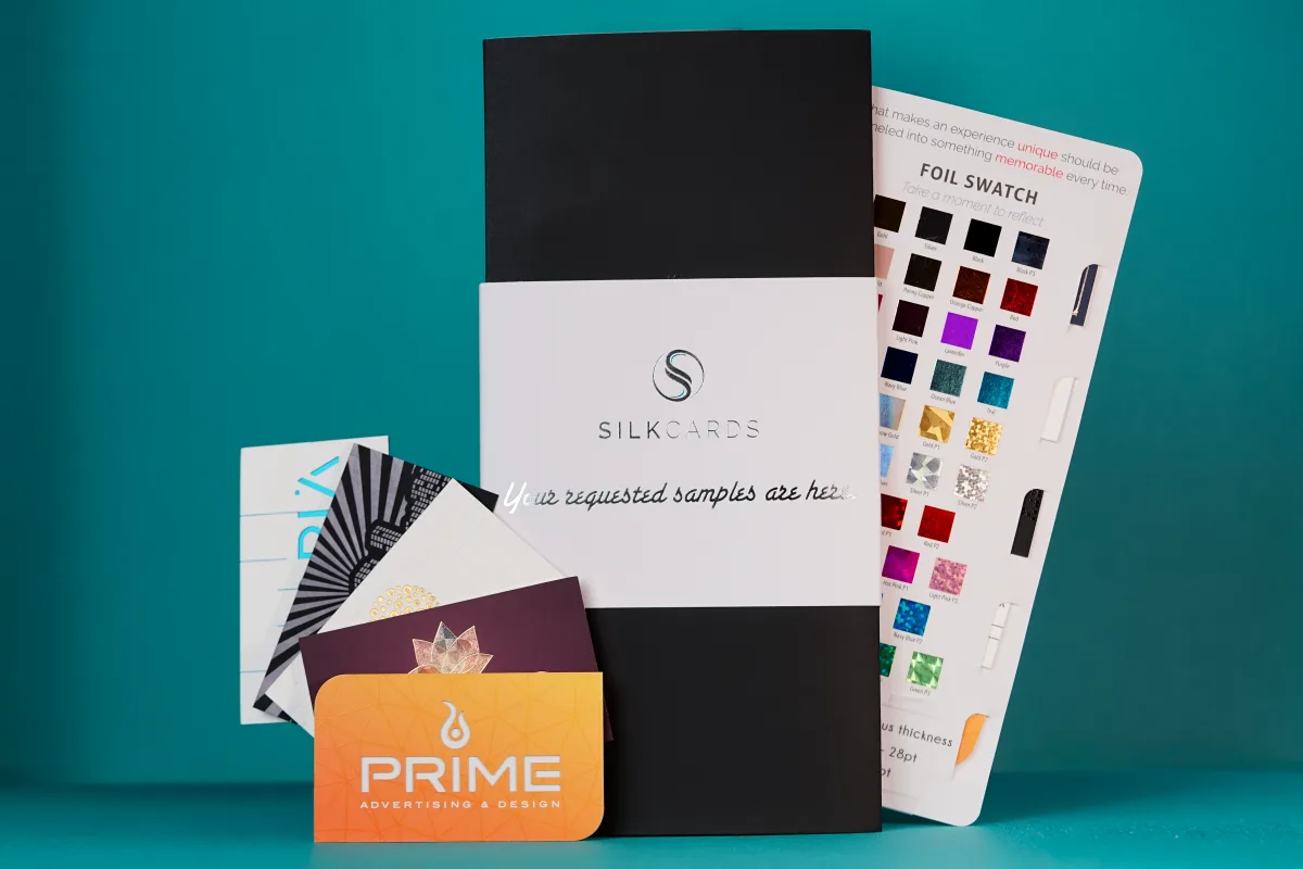 The SilkCards sample kit unboxed — a foil-stamped black and white folder, a fan of finished cards in holographic, cotton, and foil finishes, and the labeled 38-color foil swatch card tucked behind it.